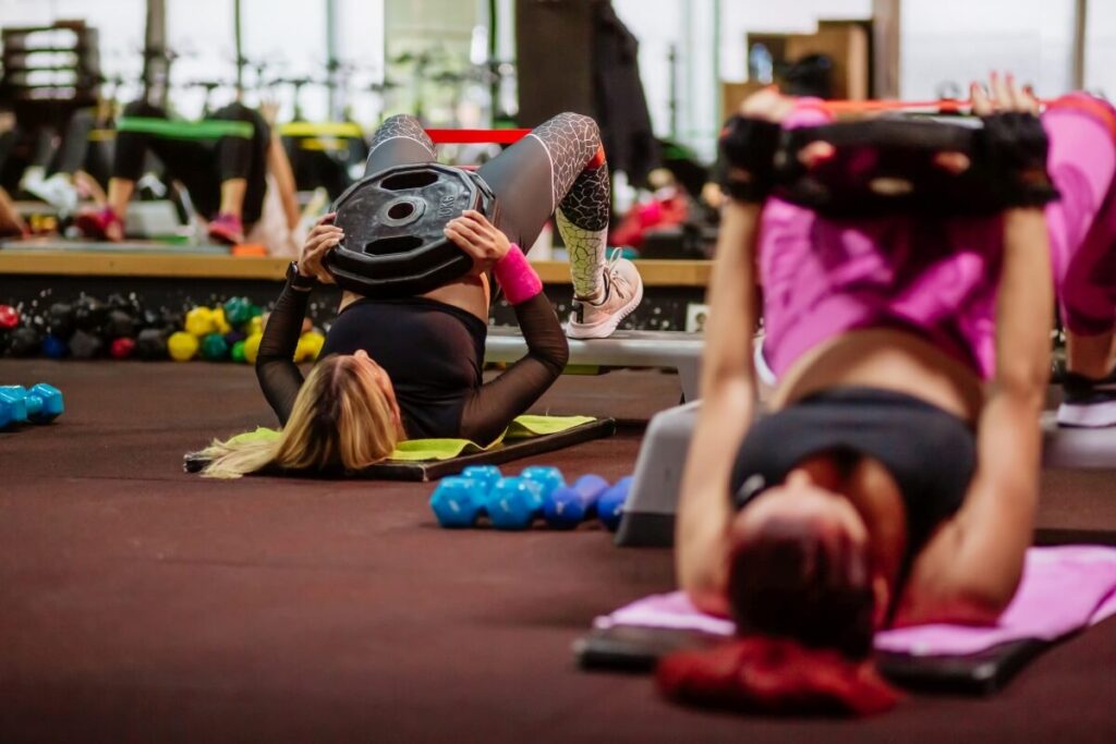 Na sali fitness uczestnicy wykonują ćwiczenia z ciężarami pod okiem trenera fitness.